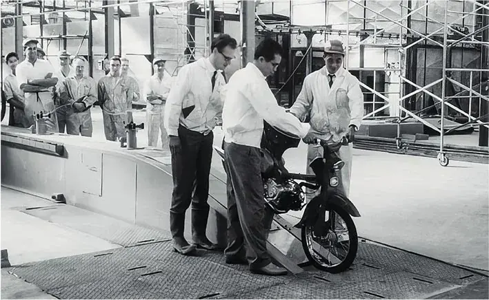 Black and white archive photograph of engineers in a factory testing a small Honda motorcycle on a rolling road, workers watching from behind