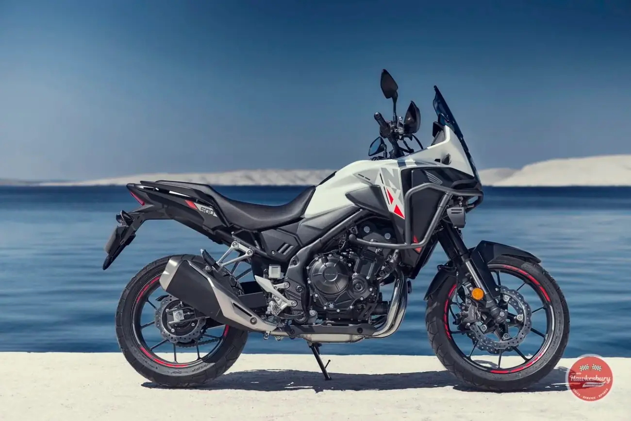 A white and black Honda NX500 adventure motorcycle parked on a sandy shoreline, calm water and pale cliffs behind