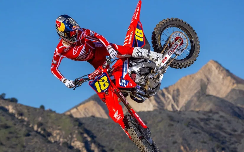 A rider in red Honda race gear whipping a red Honda CRF250 sideways in mid-air, dry mountains and blue sky behind