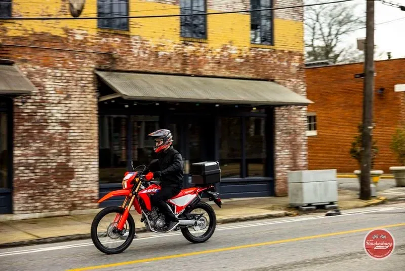 A rider in a helmet on a red and white Honda CRF300L on a city street, brick buildings behind