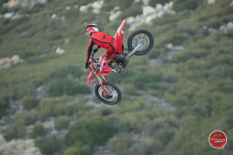 A rider in black and red gear launching a red Honda CRF450 off a dirt jump, green hills behind