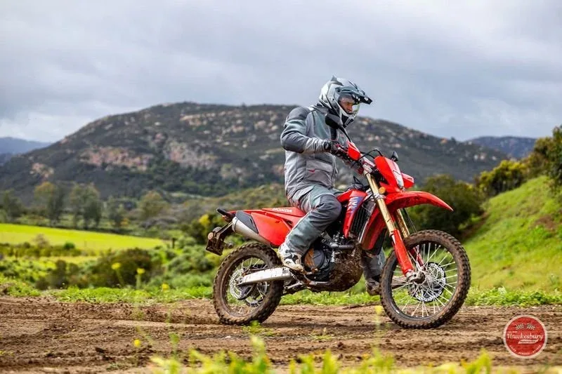 A rider in helmet and protective gear on a Honda CRF450RL on a dirt trail, green fields and mountains behind