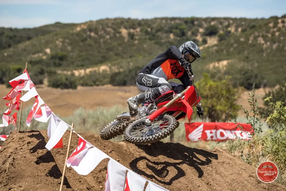 A rider on a red Honda CRF450 cresting a berm past Honda banners and course flags