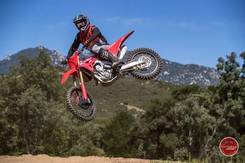 A rider in Fox gear mid-air on a red Honda CRF450 over a dirt track, mountains and blue sky behind