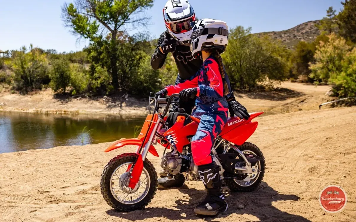A child in full riding gear on a red Honda CRF50F, an adult walking alongside