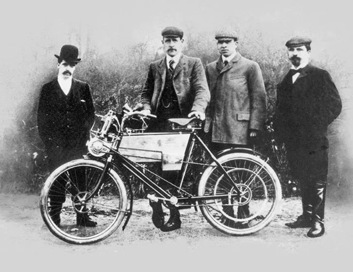 Black and white photograph of four men in period dress standing beside an early Royal Enfield motorcycle