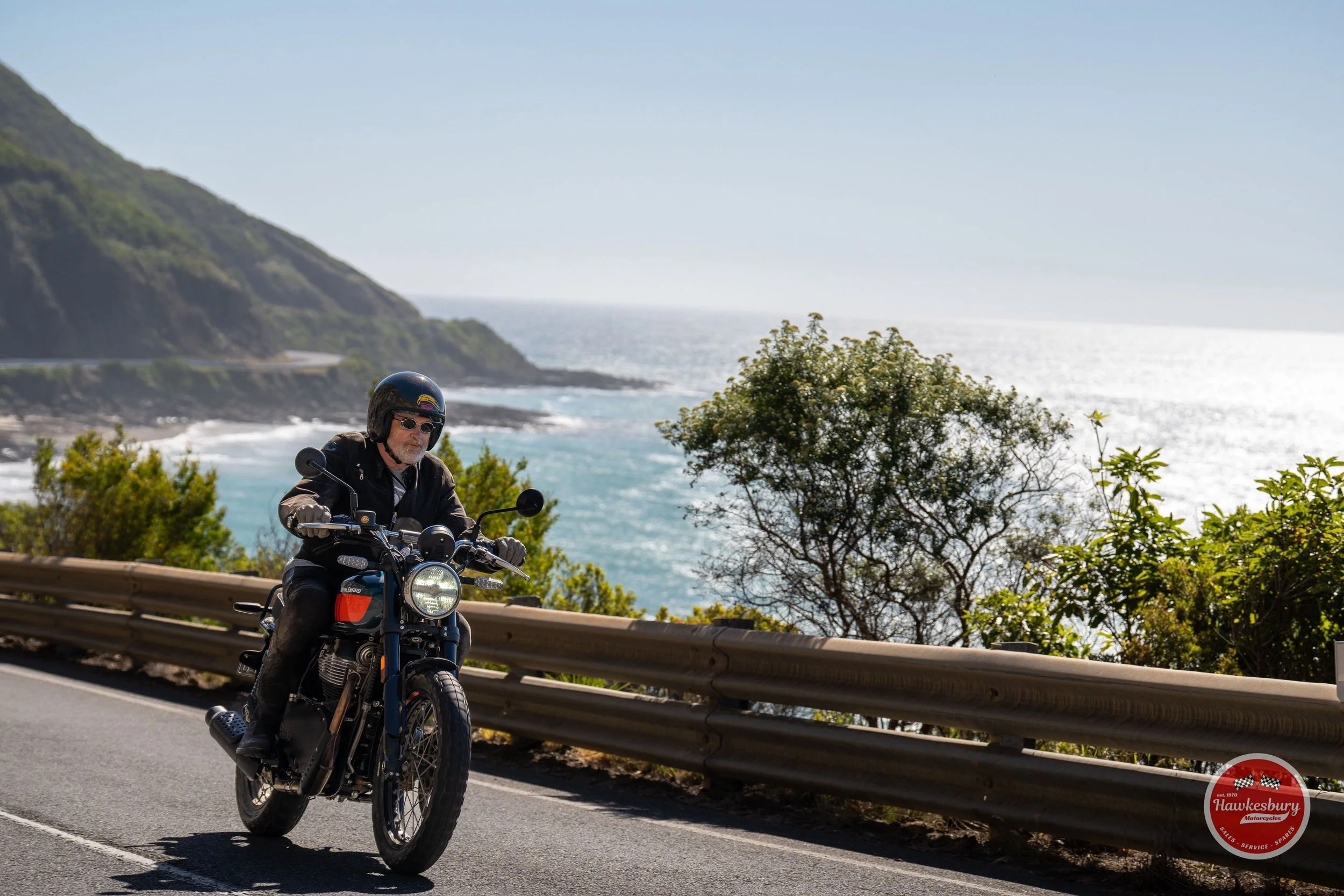 Rider on a dark Royal Enfield motorcycle on a coastal road above the ocean