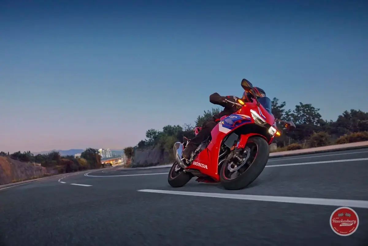 A motorcyclist in black gear riding a red and black Honda sport bike on a curving road at dusk, hills and trees behind