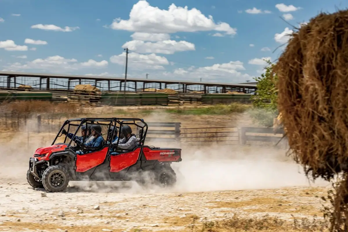 Two people driving a red Honda Pioneer Side by Side across a dusty farmyard past a round hay bale and stockyards