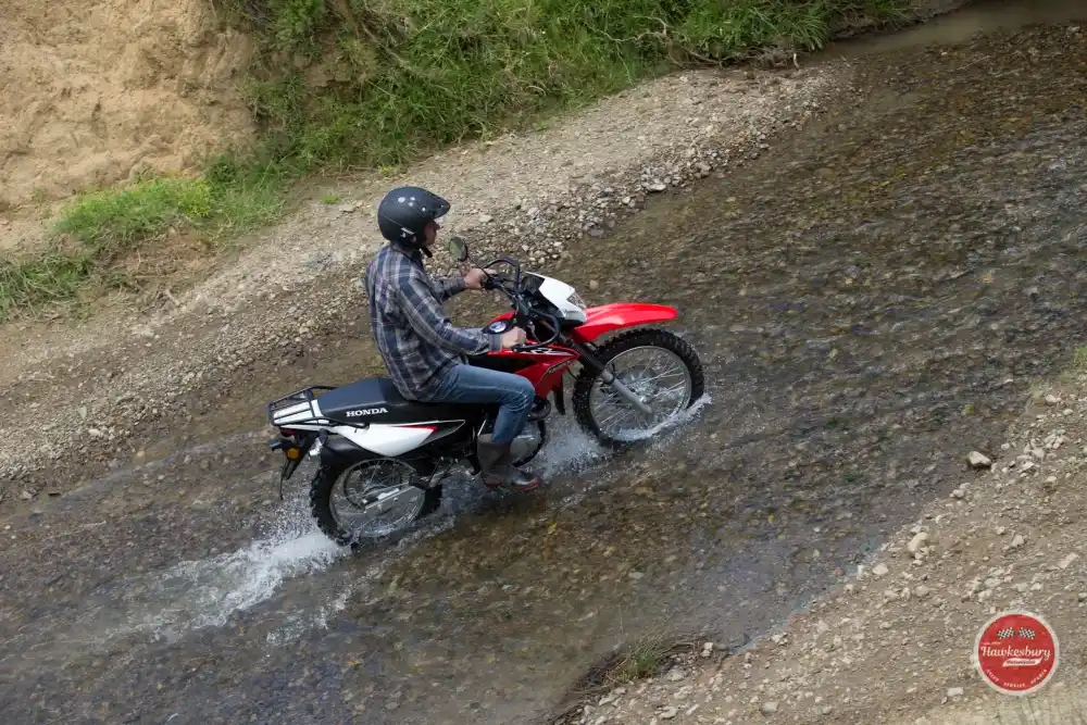 A rider on a red Honda XR150L riding through a shallow rocky creek crossing, water spraying from the front wheel. Overseas model shown.